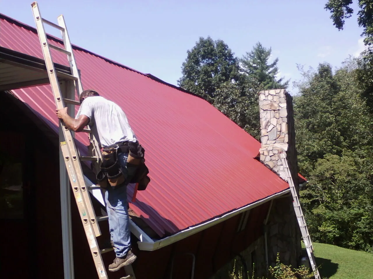 Roofer on a ladder beside a red metal roof and stone chimney — Soffit & Fascia Repair by Roofing Brothers in Olema